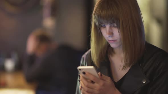 Young Beautiful Redhaired Woman Sitting in Cafe or Bar and Using a Smartphone alt