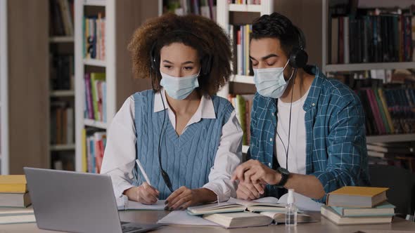 Two Students in Medical Masks Sit at Desk in University Library Listen to Teacher in Headphones on alt