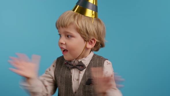 Smiling Little Birthday Boy in Cone Hat Applauding on Blue Wall Background alt