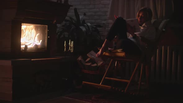 A Girl is Sitting in a Rocking Chair and Reading a Book Near the Fireplace alt