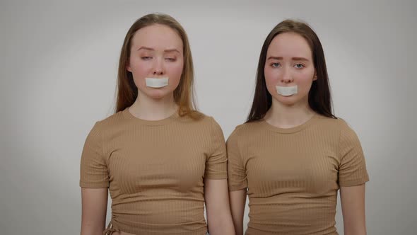 Portrait of Twin Sisters with Taped Mouth Looking at Camera with Sad Facial Expression alt