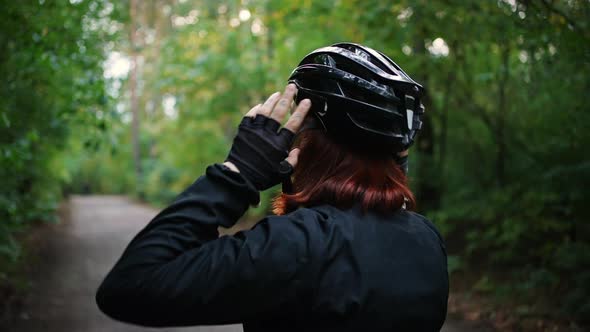 Back View Shot of Young Redhaired Woman Cyclist Putting on Protective Helmet Before Ride Sitting on alt