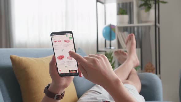 Man Lying On Couch Using Smartphone Buys In Internet Shop. Male Selects Beef In Grocery Online Store alt