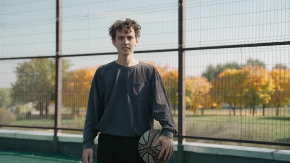 Young Guy Stands on a Basketball Court in the Park and Looks at the Camera