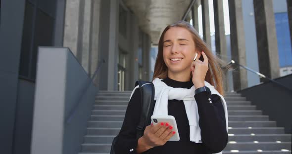 Blondie with Backpack Listening Music in Earphones while Leaving Modern city Building alt