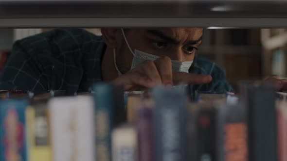 Closeup Serious Focused Young Indian Guy in Medical Mask Looking for Suitable Book in Bookstore alt