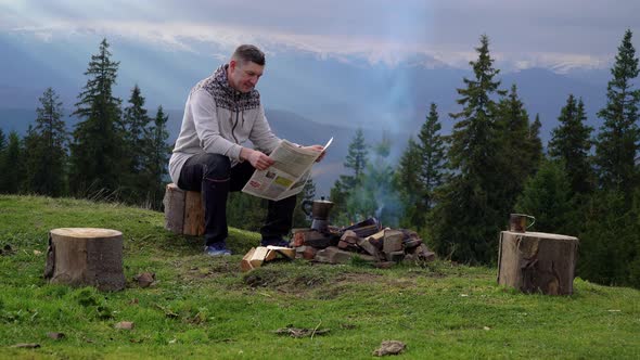 A Man Reads a Newspaper Near a Fire in the Mountains alt