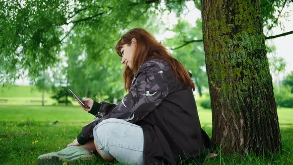 Brunette Woman Sits in the Park Under a Tree and Uses the Phone