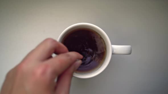Man's Hand Pours Spoonful of Sugar Into White Ceramic Mug with Tea