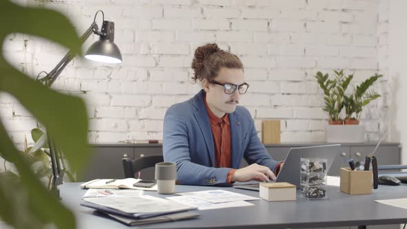 Young Hipster Businessman Working on Laptop and Picking up Ringing Smartphone alt