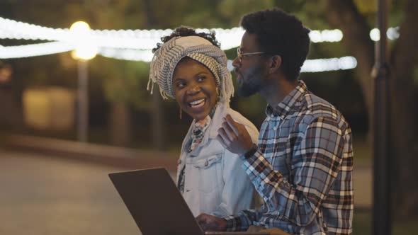 Black Man and Woman Using Laptop and Chatting in Park in Evening alt