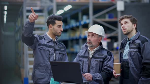 Senior Expert Man in Hard Hat Showing Logistics Plan on Laptop to Employees Talking Pointing Up at alt