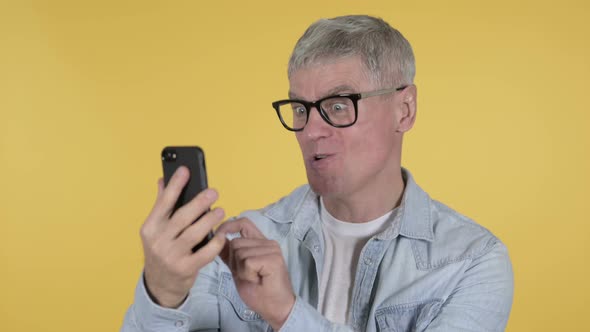 Excited Casual Senior Man Celebrating Success on Smartphone, Yellow Background alt