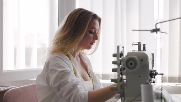 Young Beautiful Light Hair Seamstress Sews Focused in the Workplace