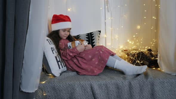 Little Cute Girl in a Santa Claus Hat Waiting for Christmas Holding a Gift While Sitting on the alt