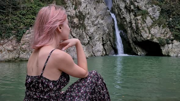 Woman in a Dress Sitting on a Stone and Enjoying a Waterfall
