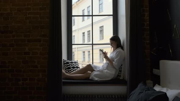 Woman in Bathrobe Using Smartphone on Windowsill alt