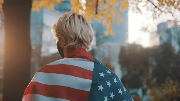 Portrait of Young Blond Woman with Face Mask Spinning with the Usa Flag Over the Shoulders alt