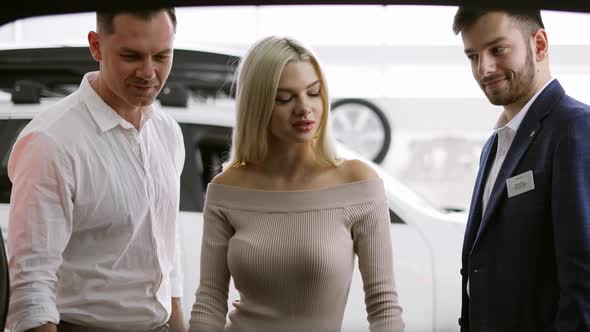 Salesman Showing the Capacity of a Trunk From Inside to Customers in Car Showroom alt
