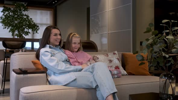 Happy young woman is sitting on cozy sofa with adorable little cute daughter. They watch TV alt
