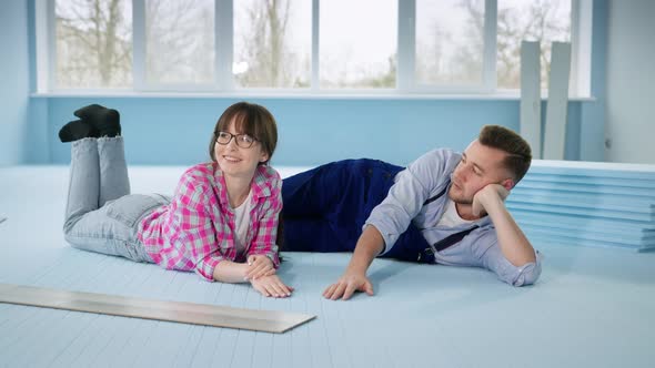 Young Couple Man and Woman Lie on Insulated Floor Discussing Laying Laminate Flooring alt