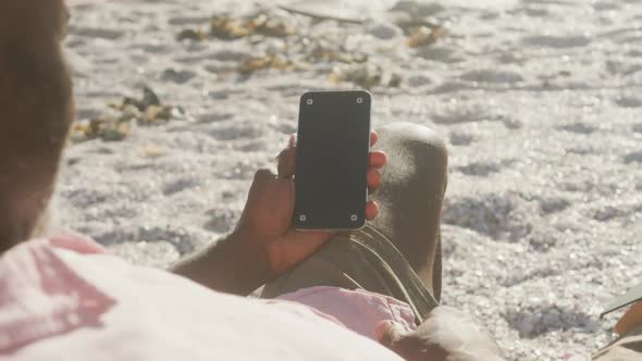 Senior african american man lying on sunbed and using smartphone on sunny beach alt