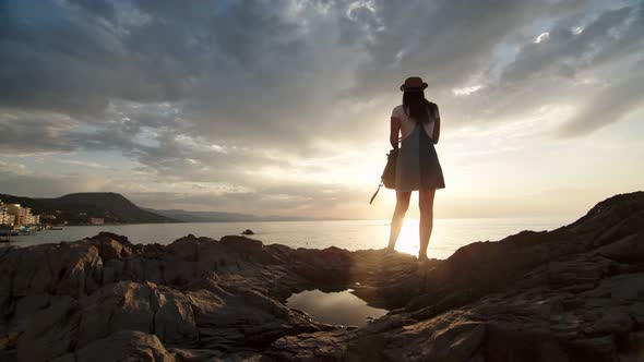 Hipster Travel Woman with Backpack Enjoying Freedom Looking on Amazing Seascape at Sunset alt