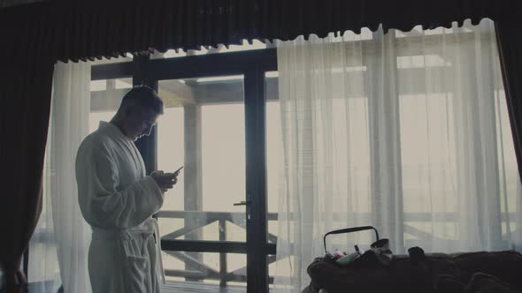 A Man In A White Robe With A Phone On The Background Of A Sunlit Terrace Of A Wooden ChaletType alt