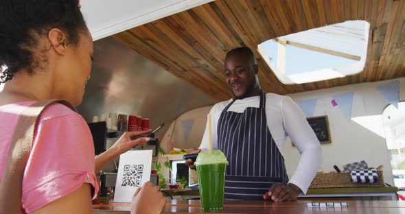 Happy female customer paying smiling african american male food truck owner by smartphone alt