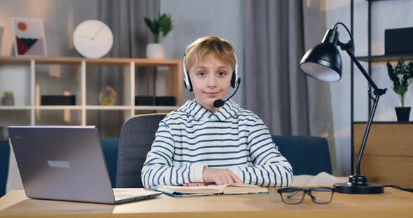 Smart Schoolboy in Headset which Sitting at his Workplace During Online Lesson and Looking at Camera alt