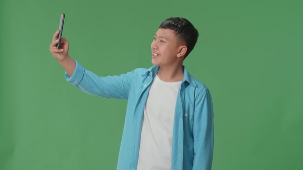 The Smiling Young Asian Boy Making Selfie While Standing On Green Screen In The Studio alt