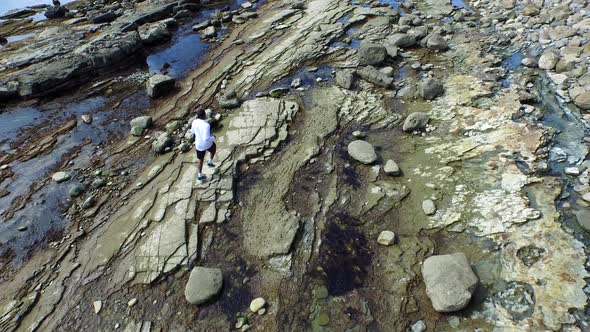 Aerial birds eye view shot of a young man running on a rocky ocean beach shoreline. alt