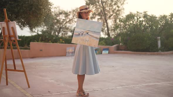 Elegant Talented Beautiful Woman Hanging Picture of Landscape on Shoulder Smiling Looking at Camera alt