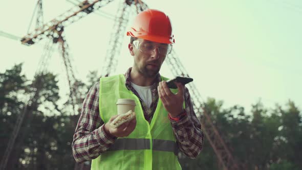 Male Worker Chief Electrician in Hard Hat Tries to Eat Sandwich and Drink Coffee but Needs to Work alt