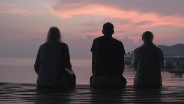 Three People Sit on the Background of the Sea in the Rays of the Setting Sun alt