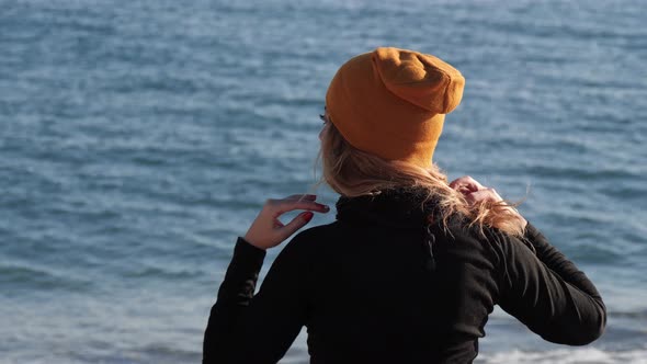 Hipster Girl in Yellow Cap Adjusting Her Blond Curly Hair in Front of Blue Sea