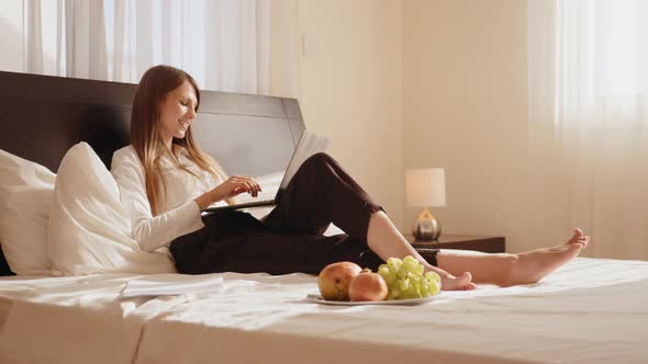 Attractive Young Woman Lying on Comfy Bed and Typing on Wireless Laptop alt