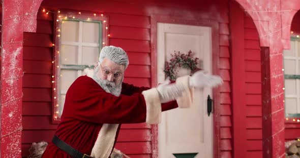 Active Santa Claus Invites Somewhere Making Hand Gestures Against the Background of a Red House with