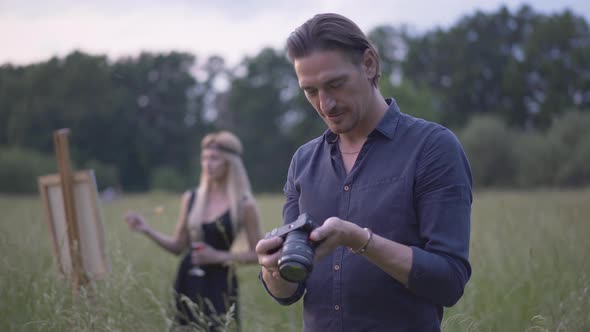 Happy Handsome Photographer Looking Through Photos on Camera As Blurred Beautiful Artist Painting at alt