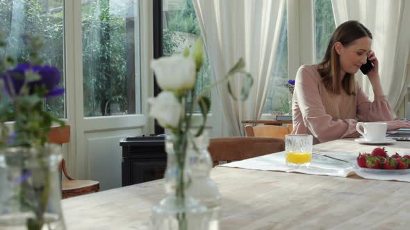 Woman at Table Talking on Phone and Working on Laptop alt