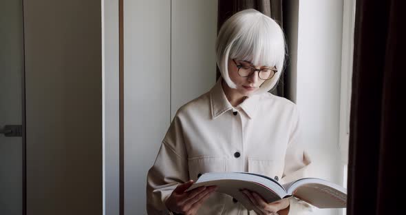 Beautiful Woman with Glasses Holds a Book in Her Hand and Looks Out the Window alt