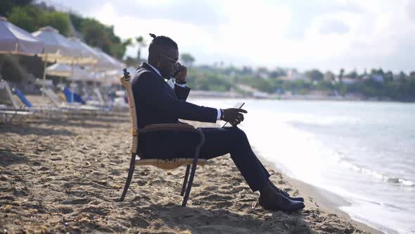 Successful Busy African American Man Talking on Smartphone Surfing Internet on Tablet Sitting on alt