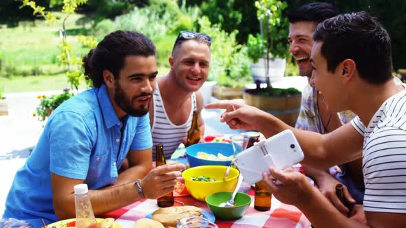 Smiling man showing his mobile phone to his friends while having meal outdoors alt