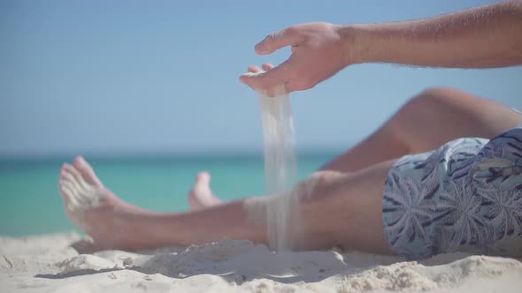 Guy Relaxing On Tropical Beach. Man Pouring Sand Through Fingers. Guy Sitting On Beach Caribbean. alt