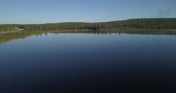 Aerial shot flying over a blue still watered lake facing a beautiful lush green forest in northern O alt