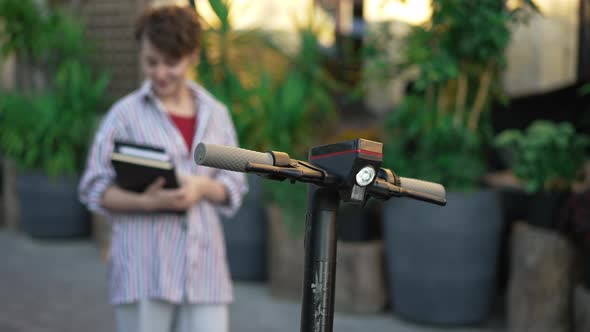 Escooter Steering Wheel with Blurred Young Slim Woman Standing at Background with Books alt