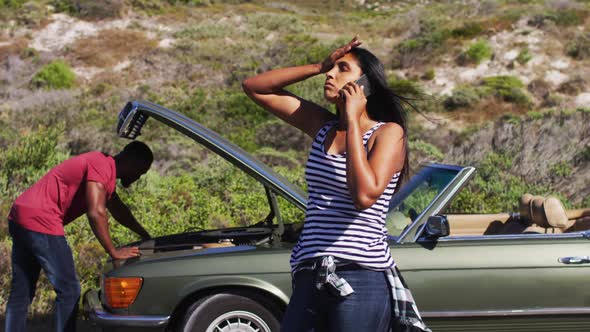 African american woman talking on smartphone while her husband trying to fix the car on road alt