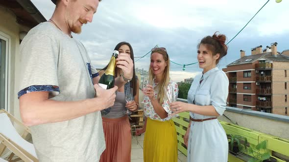 Side view of hipster man opening champagne, female friends waiting with glasses alt