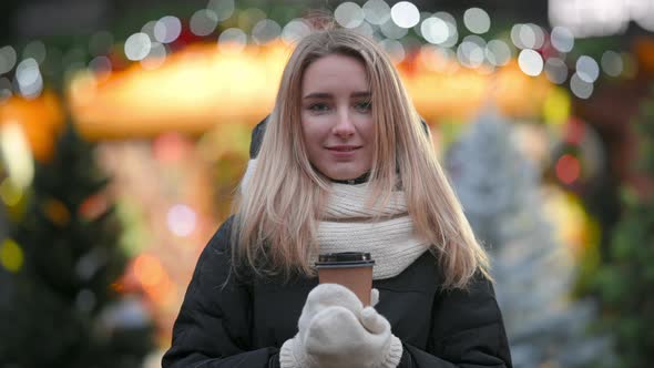 girl with a cup of hot coffee in her hands stands against the background of Christmas lights