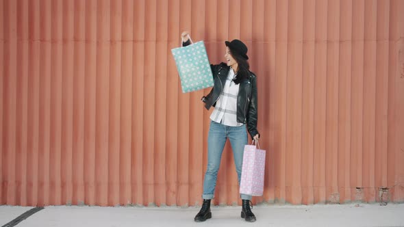 Portrait of Happy Young Lady Raising Shopping Bags and Smiling Standing Outdoors Against Wall alt